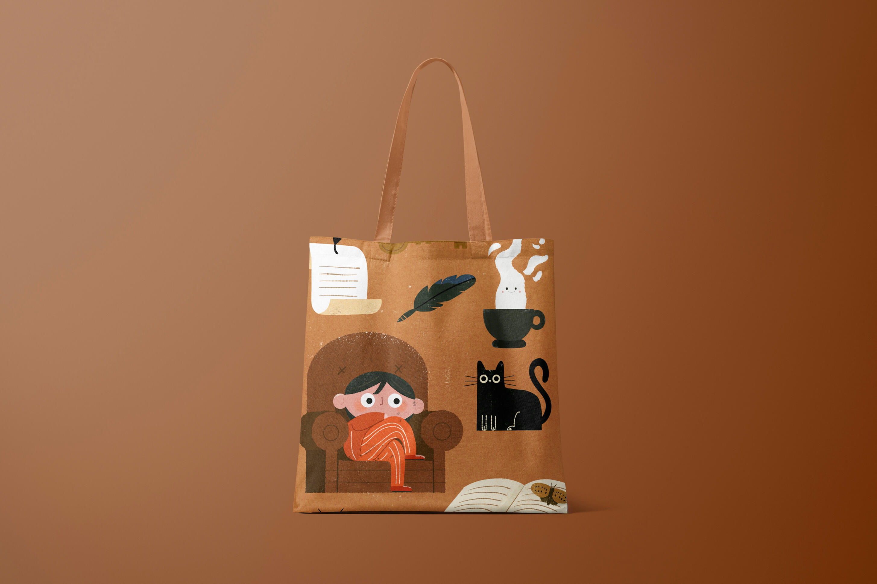 Textiles & Everyday Goods A tan canvas tote bag against a warm brown background, printed with cozy autumn illustrations: a dark-haired child wrapped in an orange blanket in an armchair, a black cat, a steaming teacup with a little ghost, a feather quill, and an open book with moth
