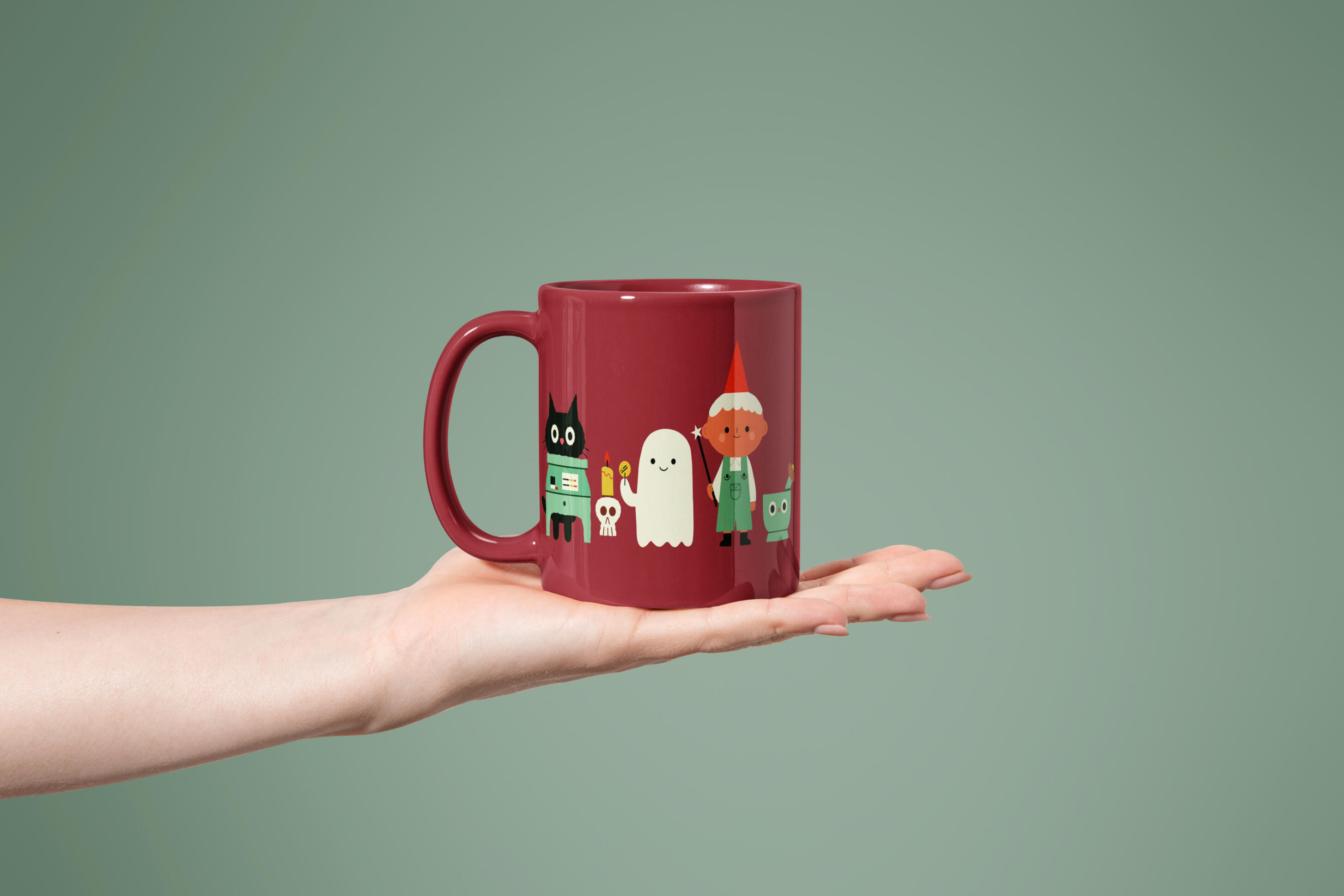 Seasonal Gifts & Ceramics A dark red ceramic mug held in an open hand against a sage green background. The mug is printed with a row of cute Halloween characters: a black cat, a small white ghost holding a candle, a child in a red pointy hat, and a skull by Prudence Moth Studio.